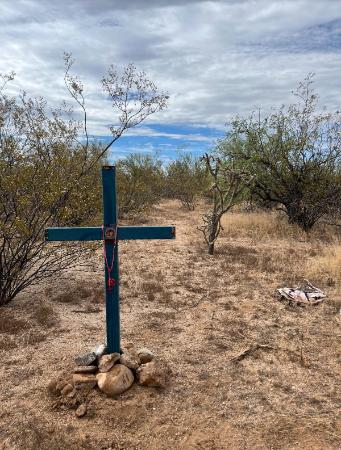 A homemade cross stands in the ground in a desert area surrounded by rocks and cacti. The location is in Southern Arizona near the U.S. - Mexico border.